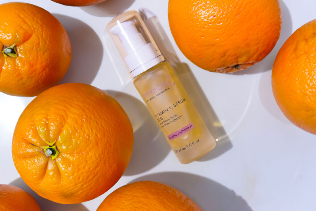 A bottle of vitamin c serum surrounded by fresh oranges on a white background, highlighting the product's citrus ingredients.