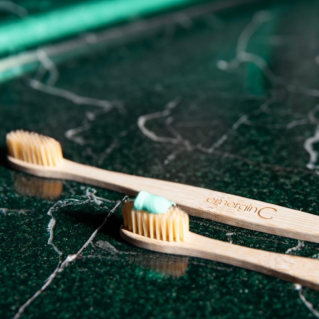 Two bamboo toothbrushes with bristles facing up, resting on a cracked green surface, with one toothbrush having a dab of pale blue toothpaste on it, suggesting a sustainable and environmentally friendly oral hygiene routine.