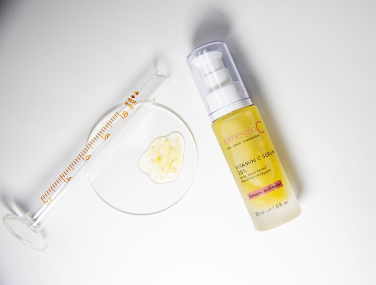 A bottle of emerginc vitamin c serum alongside a glass scientific measuring tool with a dollop of a creamy substance, against a white background, depicting a blend of skincare and science.