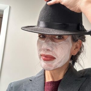 A person with a facial mask playfully tipping a black hat while posing for a selfie.