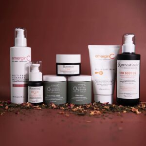 A collection of emerginc and rawceuticals skin care products elegantly displayed against a rich burgundy background with scattered botanical elements.