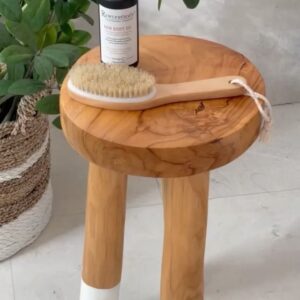 A body brush rests on top of a wooden stool with natural grain patterns, beside a bottle of skincare oil, giving a sense of a calming, organic self-care routine in a minimalist setting with a touch of greenery from a potted plant.