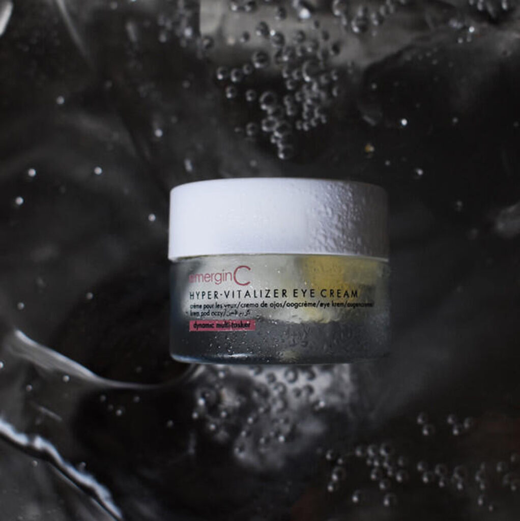 A jar of emerginc hyper-vitalizer eye cream resting on a glossy, wet surface with water droplets scattered around, conveying a sense of hydration and skincare.