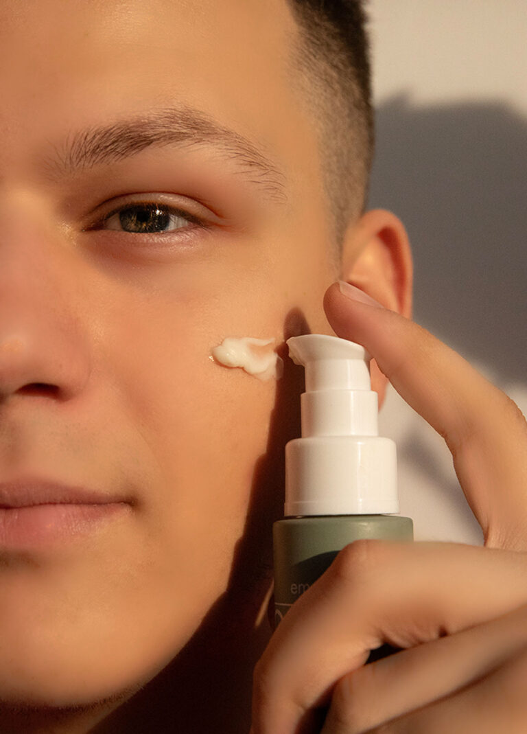 A person applying a dab of cream from a pump bottle onto their cheek, with sunlight casting soft shadows on the face, highlighting the skincare routine.