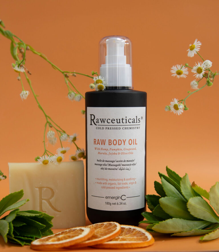 A bottle of rawceuticals cold-pressed body oil with a beige soap bar, surrounded by natural elements like sliced dried oranges and white daisy flowers against an earth-toned background.