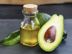 A bottle of avocado oil with a ripe avocado cut in half, displaying its pit, alongside fresh green leaves, presented on a wooden surface.