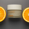 A jar of EmerginC vitamin C + retinol mask flanked by two vibrant orange slices on a dark background, depicting a theme of natural radiance and skin care.
