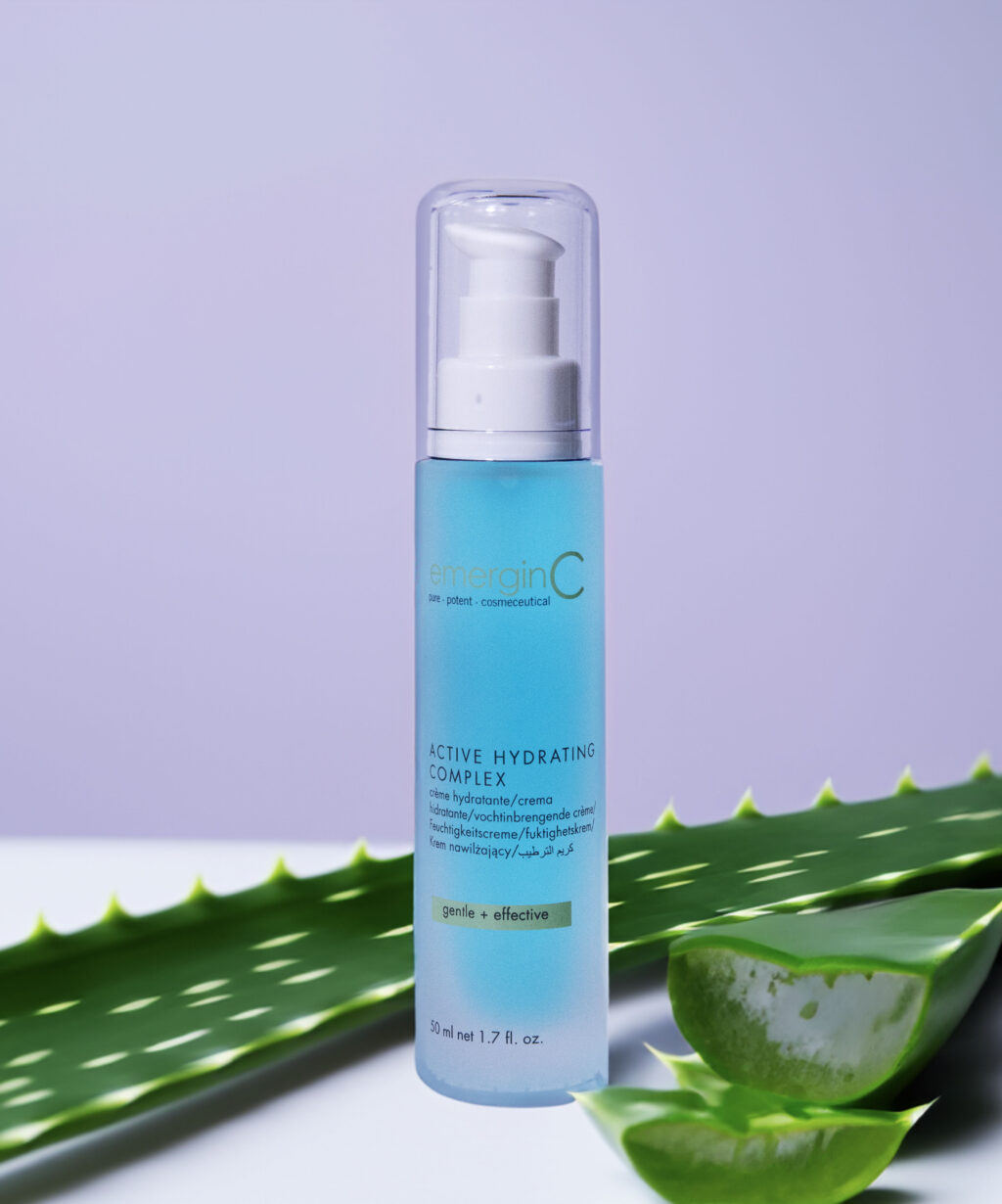 A sleek bottle of emerginc active hydrating complex skincare product positioned in front of fresh aloe vera leaves on a soft purple background, symbolizing gentle and effective hydration care.
