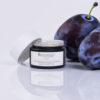 A jar of rawceuticals' BLEND™ skincare product elegantly presented beside two fresh, ripe plums on a clean, white surface, highlighting the theme of natural ingredients.