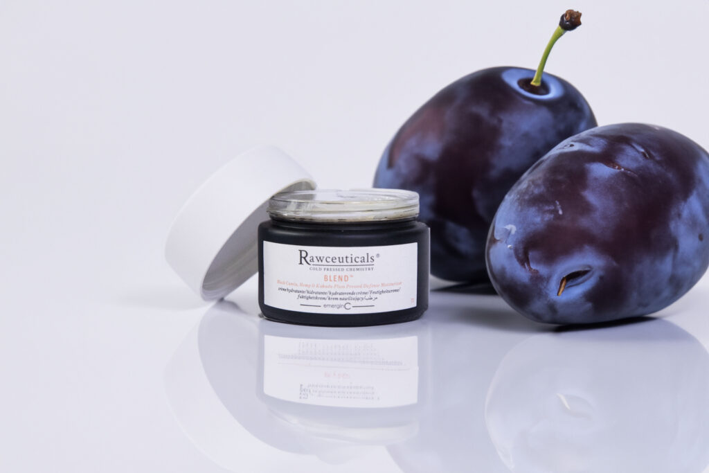 A jar of rawceuticals' BLEND™ skincare product elegantly presented beside two fresh, ripe plums on a clean, white surface, highlighting the theme of natural ingredients.