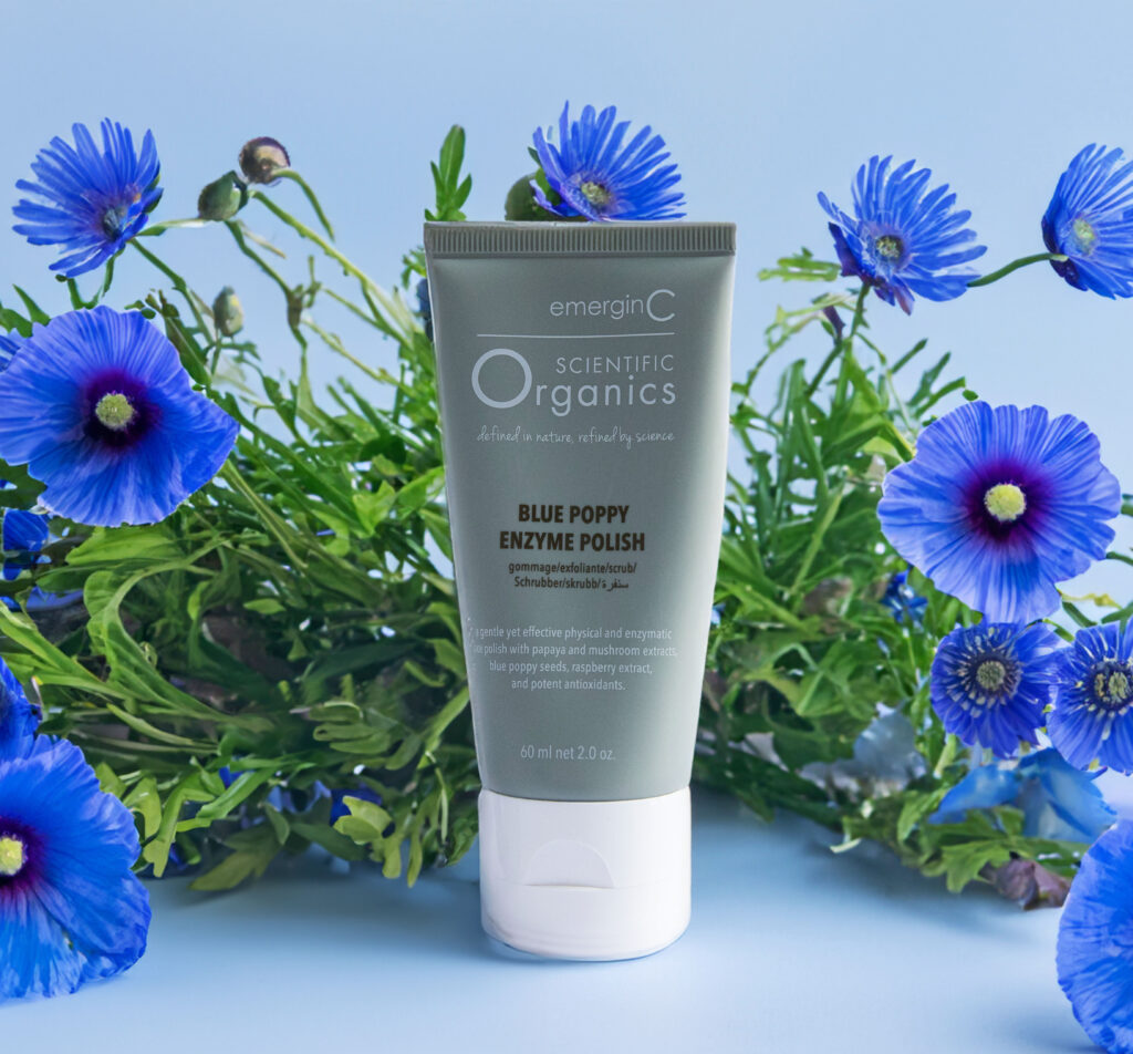 A tube of emerginc scientific organics Blue Poppy Enzyme Polish surrounded by vibrant blue poppy flowers against a calming blue background.