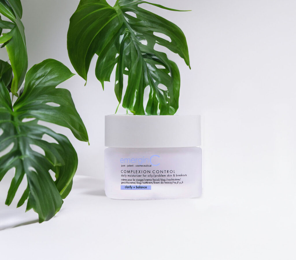 A skin care product from emerginc labeled "Revive Complexion Control" is prominently displayed in a clean, minimalist setting, flanked by vibrant green leaves that suggest a natural or botanical aspect to the product.