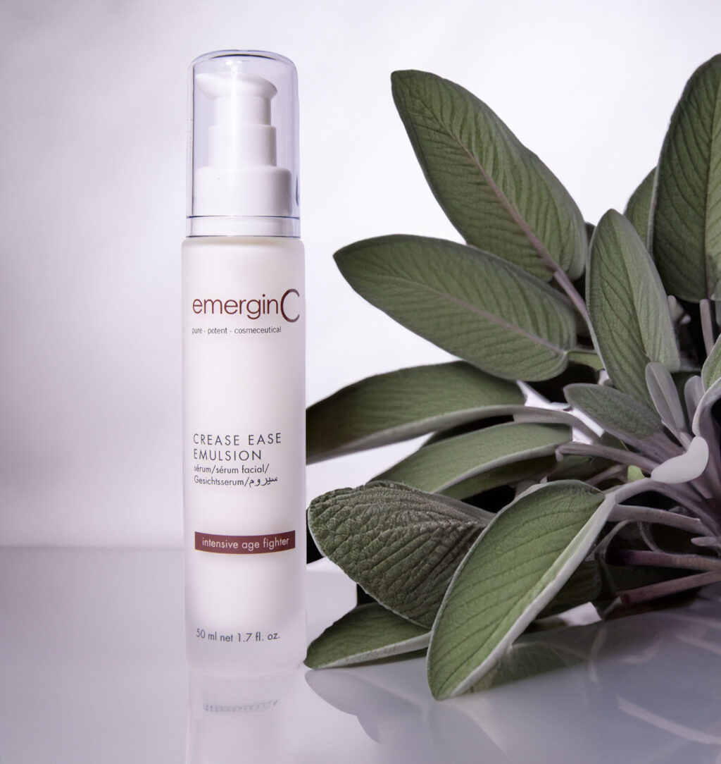 Sentence with product name: A bottle of emerginc crease ease emulsion stands prominently in the foreground with fresh green leaves artistically arranged in the background, all against a clean, white backdrop, evoking a sense of natural skincare.