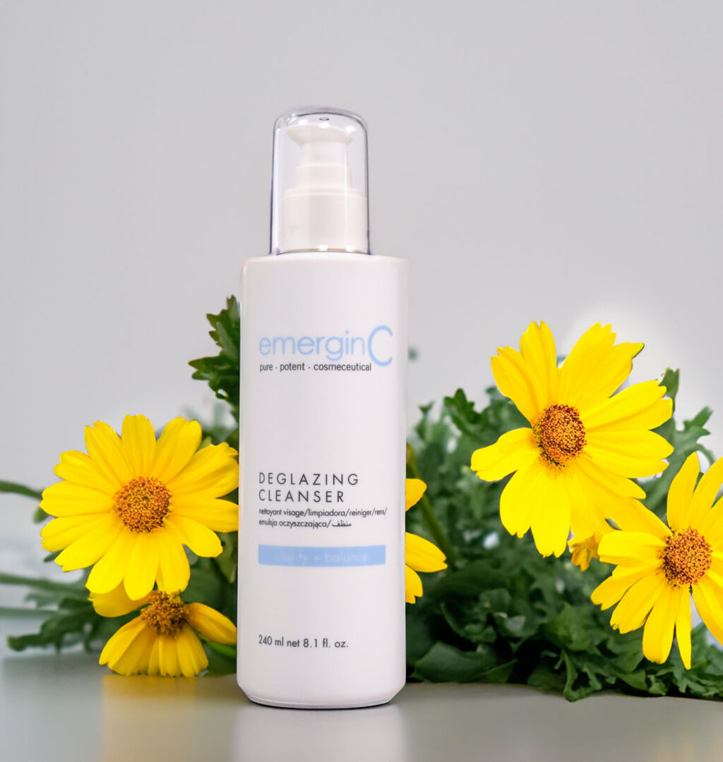 A bottle of emerginc deglazing cleanser flanked by vibrant yellow flowers against a neutral background.
