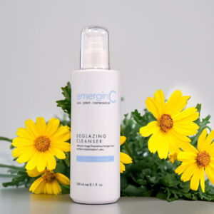 A bottle of emerginc deglazing cleanser flanked by vibrant yellow flowers against a neutral background.