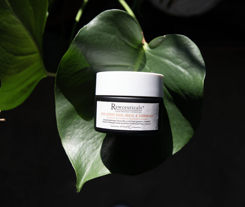Jar of Red Berry Dual Mask & Gommage™ nestled in the embrace of vibrant green leaves, illuminated by a beam of sunlight.