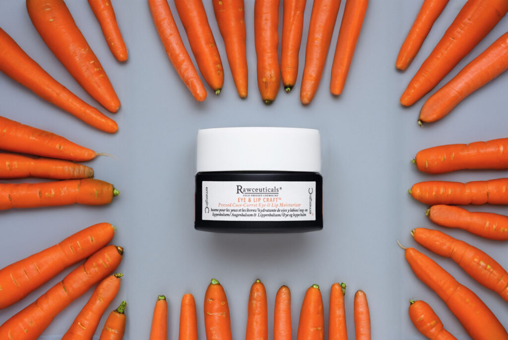 A jar of EYE & LIP CRAFT™ surrounded by a circle of fresh carrots on a blue background, suggesting the product contains carrot extracts or vitamins beneficial for the skin.