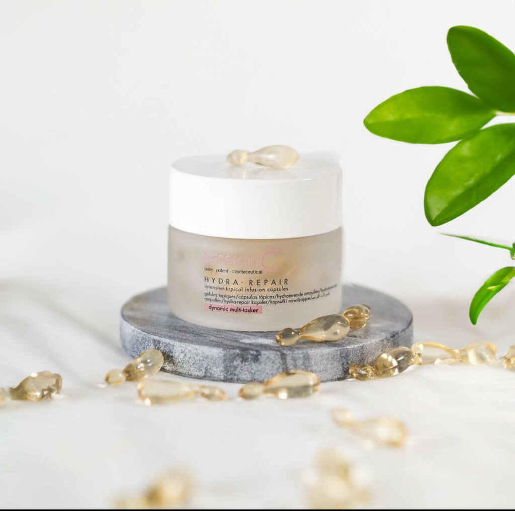 A jar of hydra-repair intensive topical infusion capsules placed on a marble stand, surrounded by a string of pearls and a green plant, against a clean, white background.