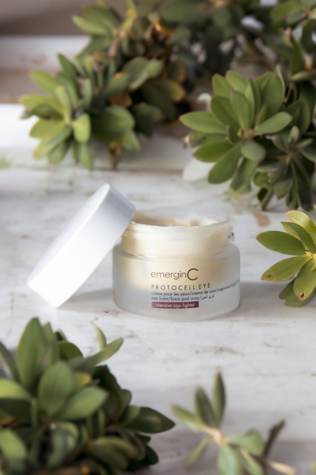 A jar of EmerginC Protocell Eye Cream surrounded by fresh green succulents on a marble surface, suggesting a natural and luxurious skincare routine.