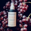 A bottle of RAW BODY OIL surrounded by clusters of red grapes against a dark background.