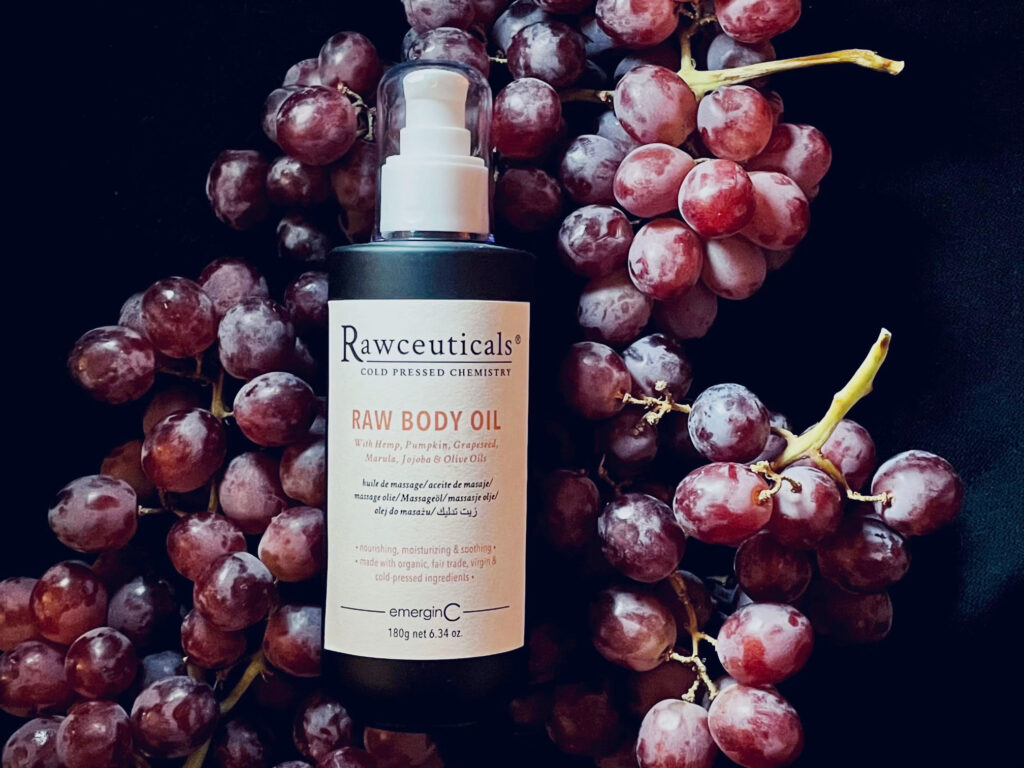 A bottle of RAW BODY OIL surrounded by clusters of red grapes against a dark background.