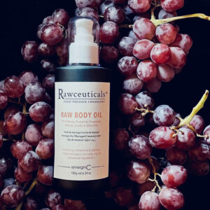 A bottle of RAW BODY OIL surrounded by clusters of red grapes against a dark background.