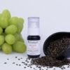 A bottle of RAW FACIAL OIL™ next to a bunch of green grapes with a scoop of scattered flaxseeds on a white background.