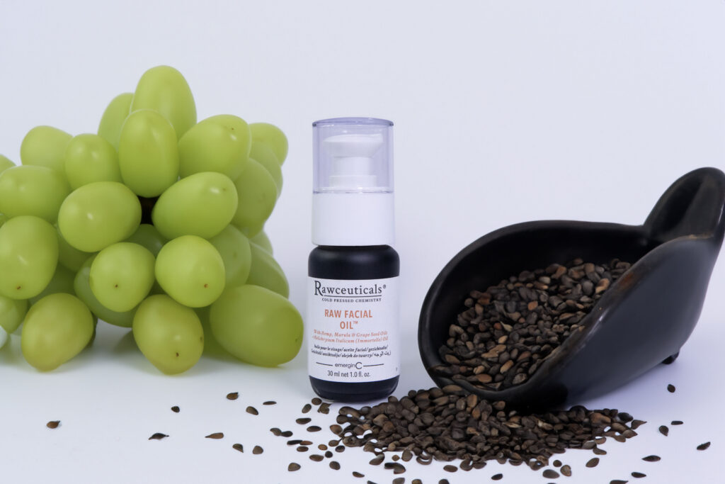A bottle of RAW FACIAL OIL™ next to a bunch of green grapes with a scoop of scattered flaxseeds on a white background.