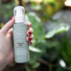 A hand with red nail polish holds a bottle of Spirulina Toner against a backdrop of verdant plants.