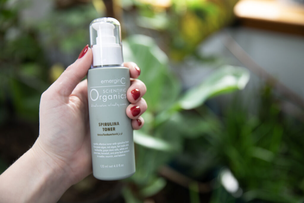 A hand with red nail polish holds a bottle of Spirulina Toner against a backdrop of verdant plants.