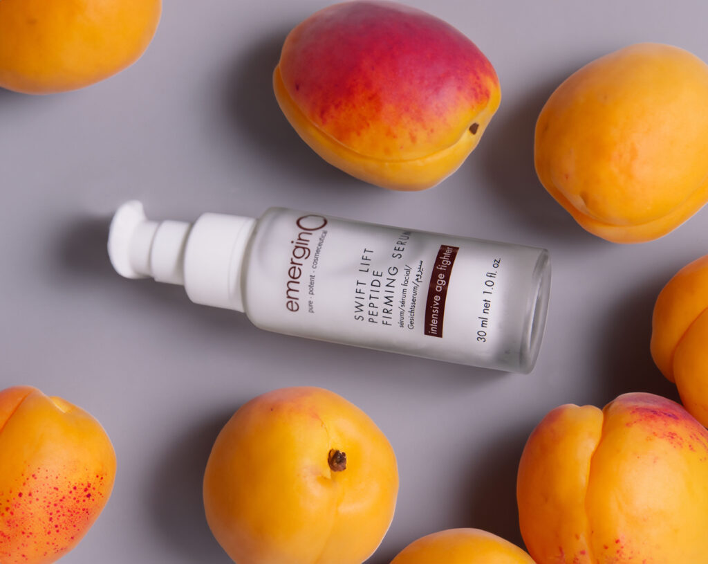 Bottle of swift lift® peptide firming serum surrounded by fresh peaches on a gray surface.