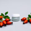 A jar of eyelight cream flanked by fresh rose hips on a neutral background, suggesting natural ingredients in cosmetics.