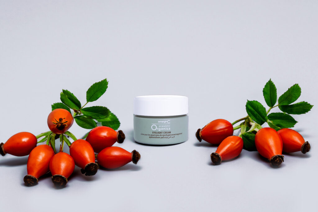 A jar of eyelight cream flanked by fresh rose hips on a neutral background, suggesting natural ingredients in cosmetics.