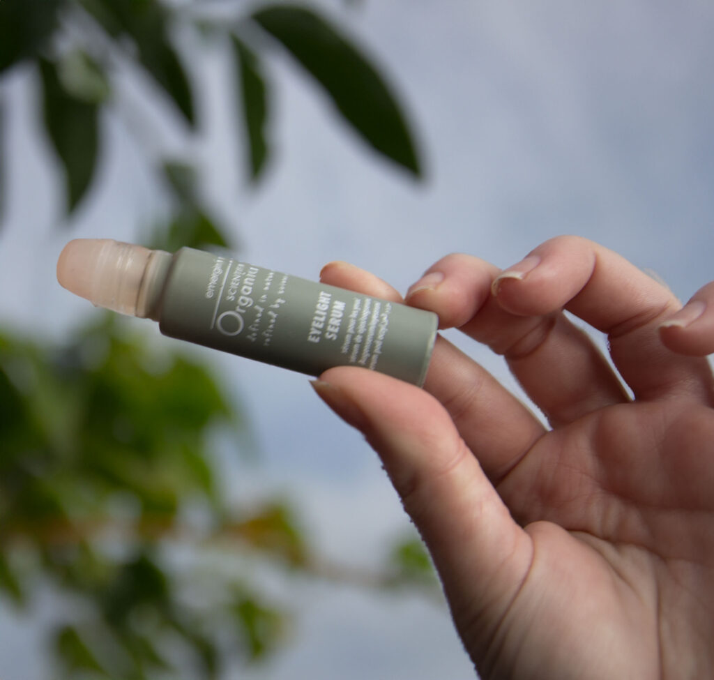 A hand holding a small tube of eyelight serum with a blurred natural background, suggesting a connection to nature and organic ingredients.