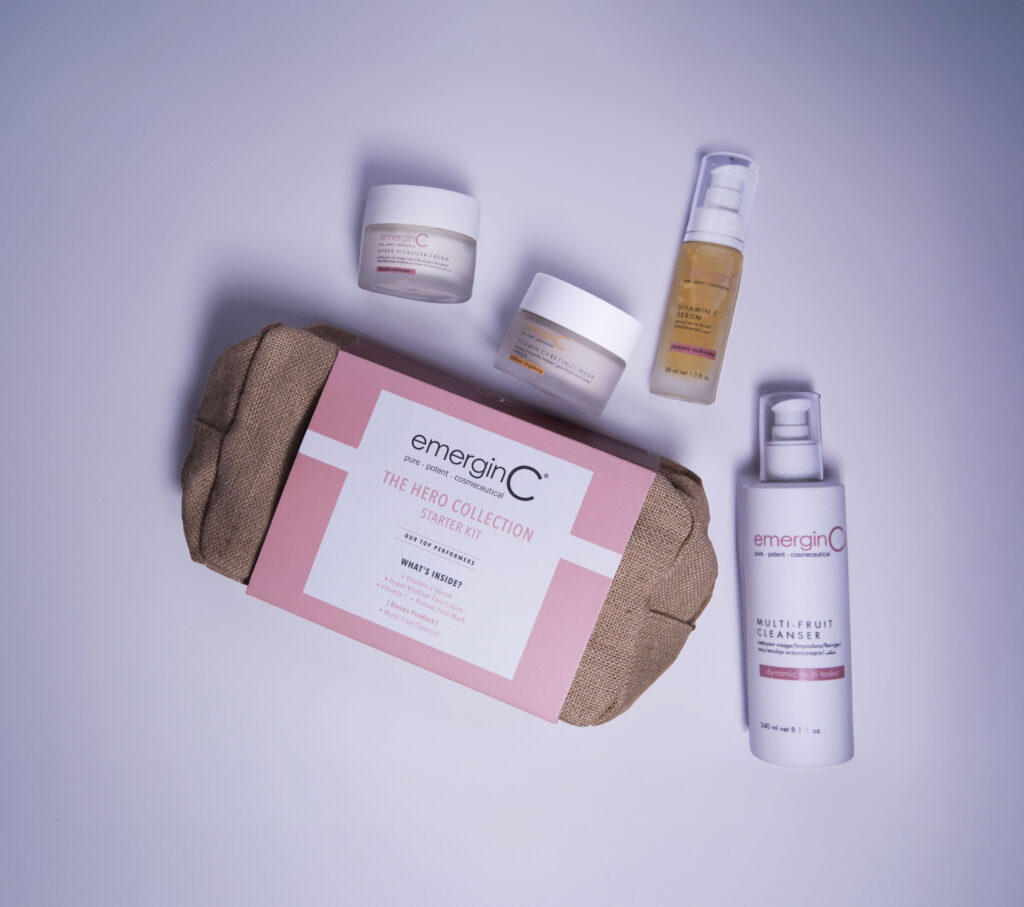A set of EmerginC skincare products arranged neatly, featuring The Hero Collection Starter Kit, alongside a multi-fruit cleanser and other skin treatment jars, against a pale lavender background.