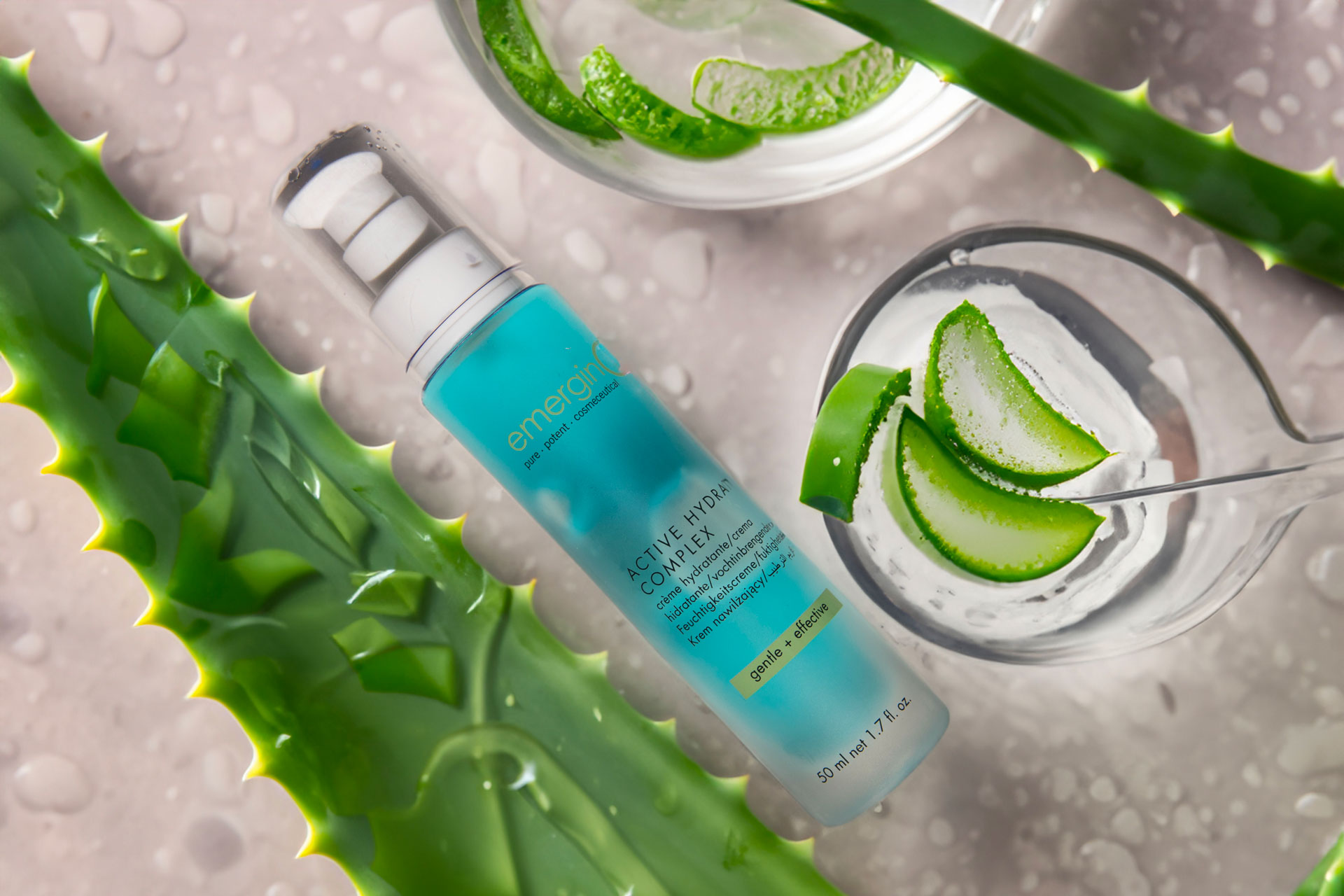 A skincare product bottle labeled "energizing comfort" surrounded by fresh aloe vera pieces, lime slices, and a glass of water, set on a reflective watery surface.