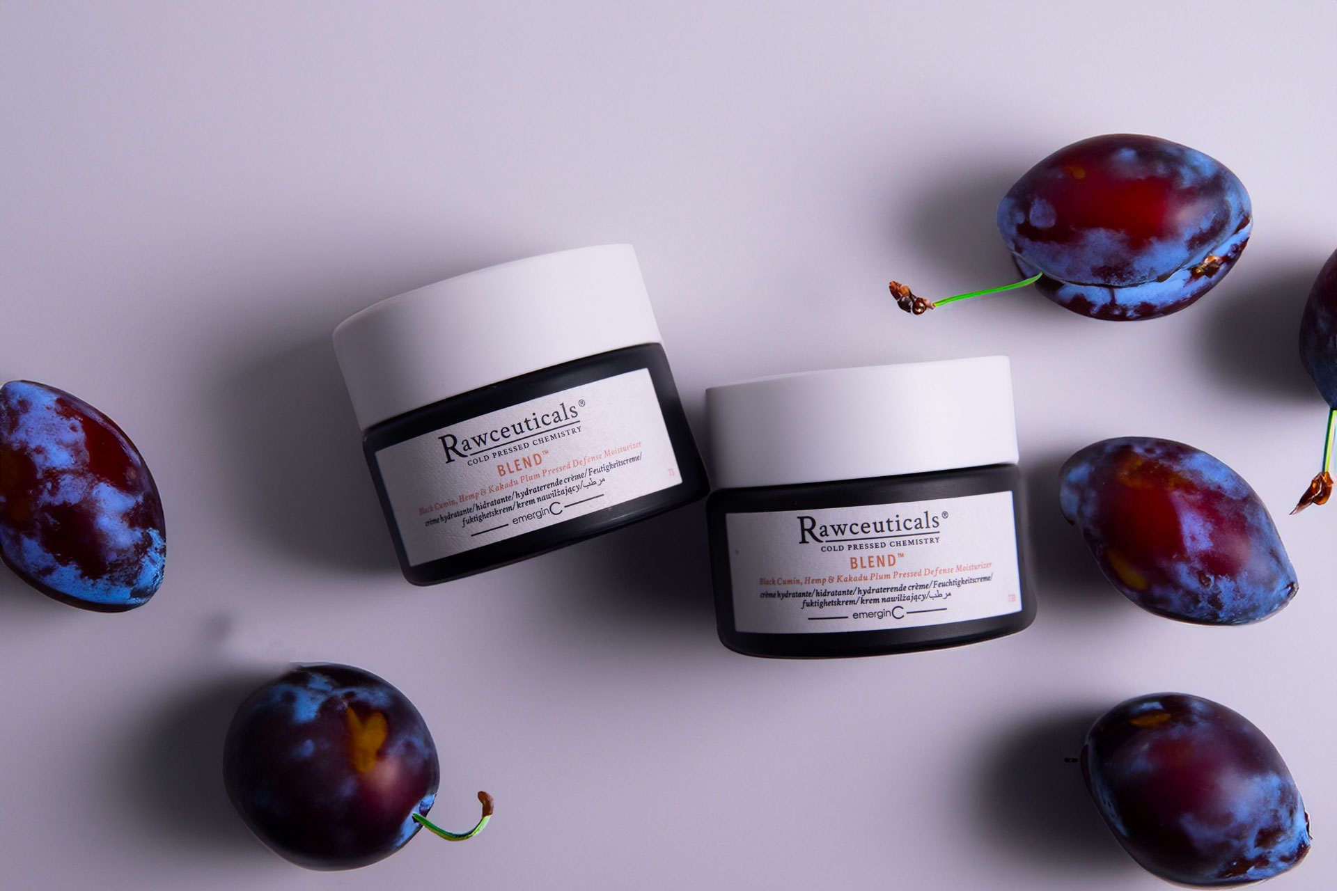 Two jars of rawceuticals skincare products surrounded by fresh plums on a light grey background.