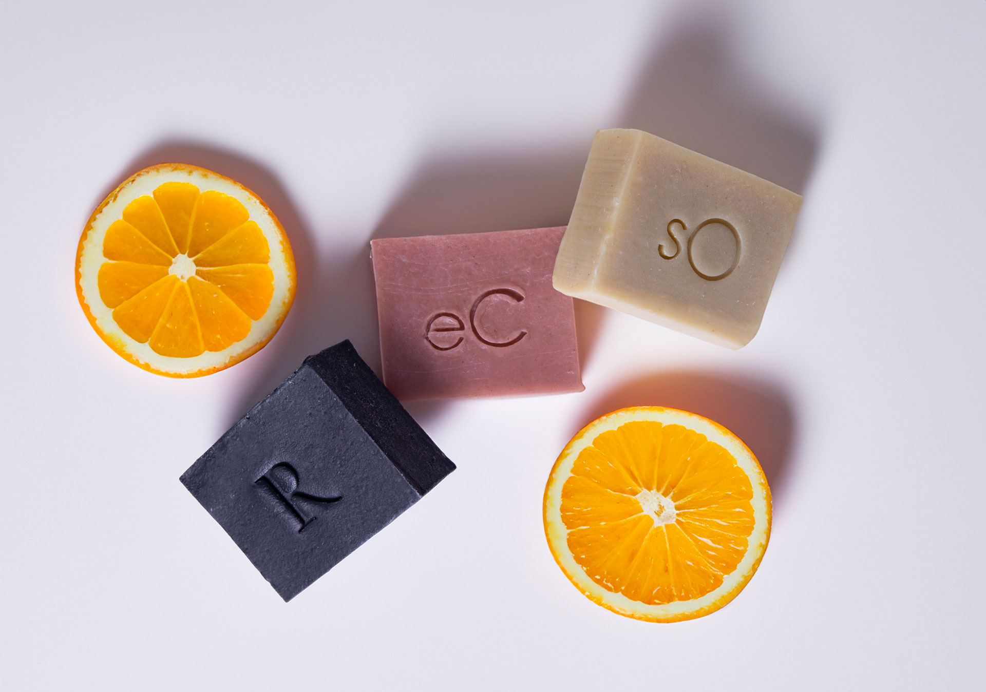 Four colorful bars of soap labeled ec, so, and r arranged with two orange slices on a light background, showcasing a vibrant and clean aesthetic.