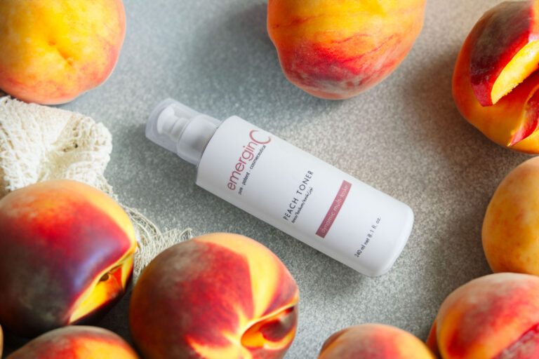 A bottle of emerginc vitamin c serum placed on a table surrounded by fresh, ripe peaches. the setting has a natural, wholesome look with a focus on the skincare product among the fruit.