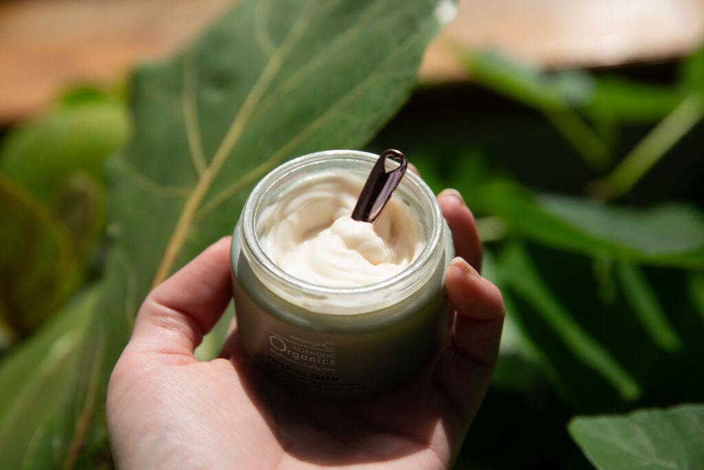 A hand holding an open jar of organic face cream with a small spatula on top, surrounded by green leaves in a natural setting.