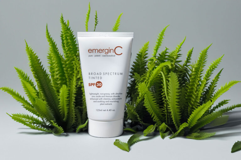 A tube of emerginc tinted sunscreen spf 30 surrounded by lush green ferns against a light gray background.