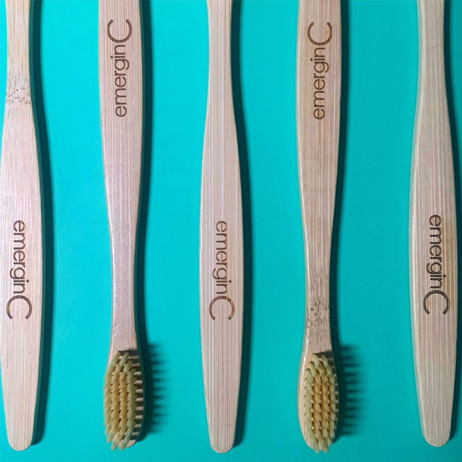 Four bamboo toothbrushes with green bristles, aligned horizontally, displayed on a turquoise background. each brush is marked with the brand 