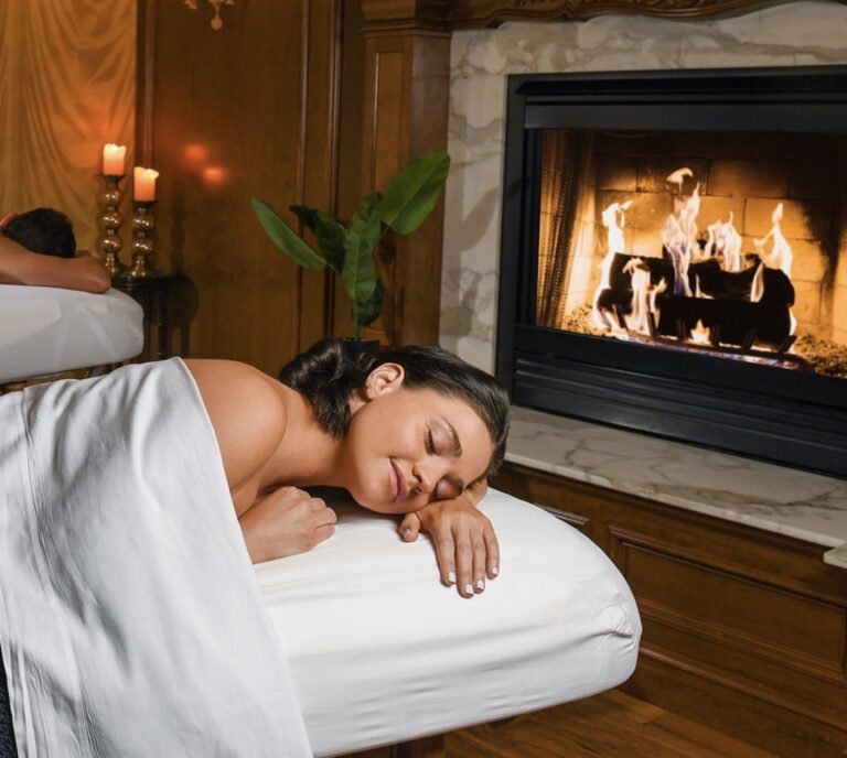 A woman relaxes on a massage table with a white sheet draped over her, eyes closed, near a lit fireplace in a cozy, candle-lit spa room with wood paneling and elegant decor.