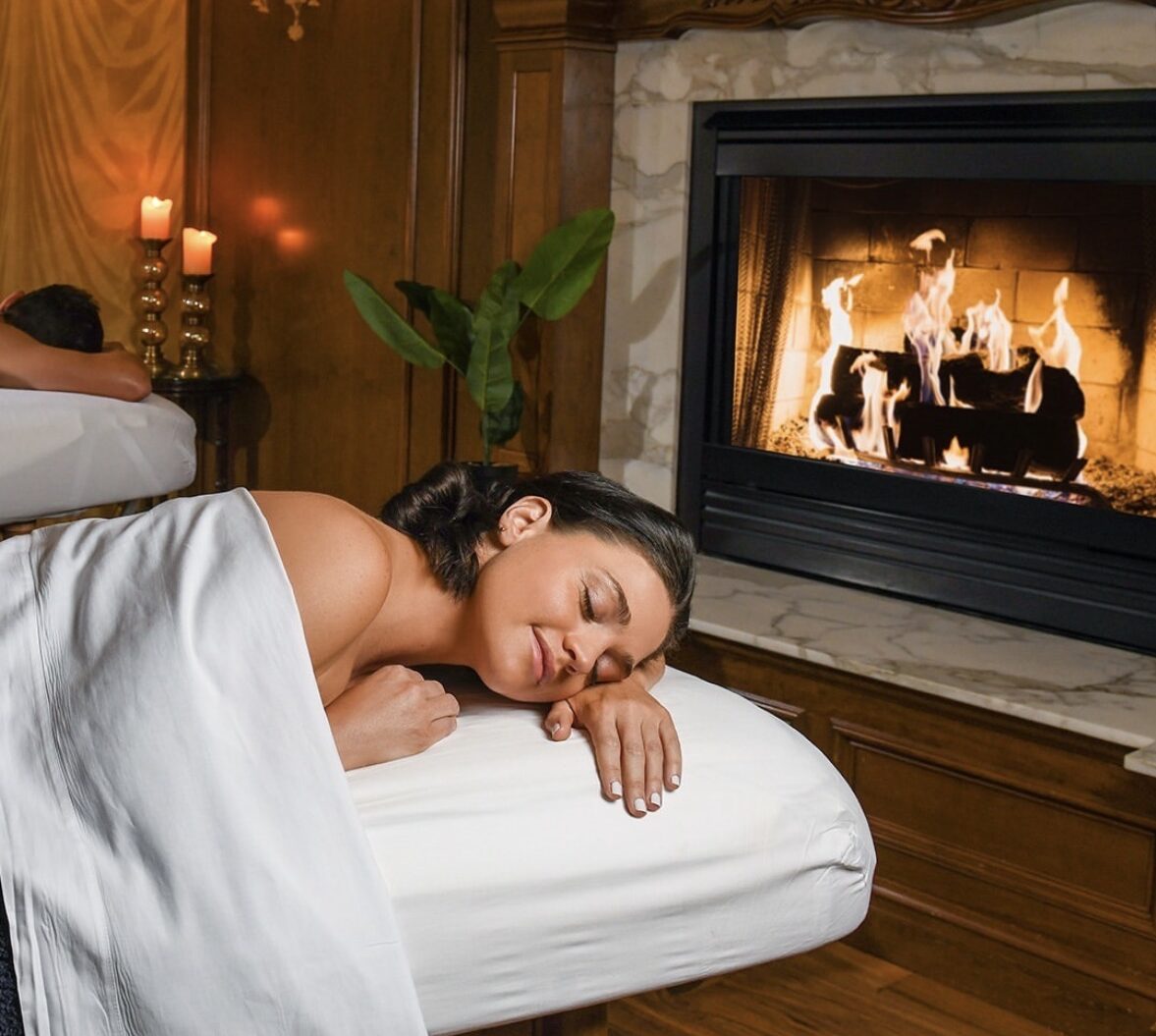 A woman relaxes on a massage table with a white sheet draped over her, eyes closed, near a lit fireplace in a cozy, candle-lit spa room with wood paneling and elegant decor.