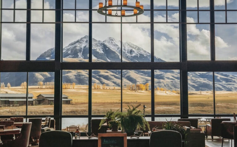 Large floor-to-ceiling windows frame a scenic view of snow-capped mountains, grassy fields, and a few small buildings. Indoors, cozy lounge seating and plants create a warm, inviting atmosphere.