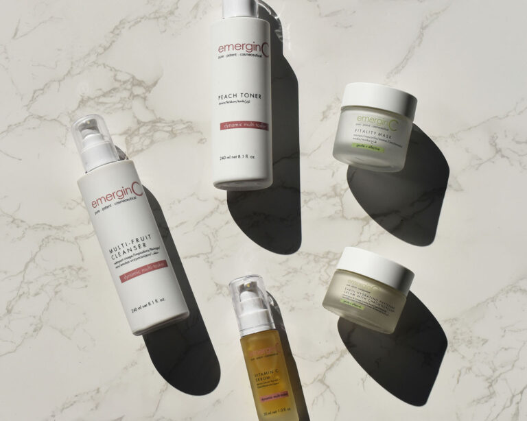 Five EmerginC skin care lines for spas—Multi-Fruit Cleanser, Peach Toner, Vitality Mask, a moisturizer, and a Vitamin C serum—are arranged on a marble surface in bright sunlight, casting striking shadows.