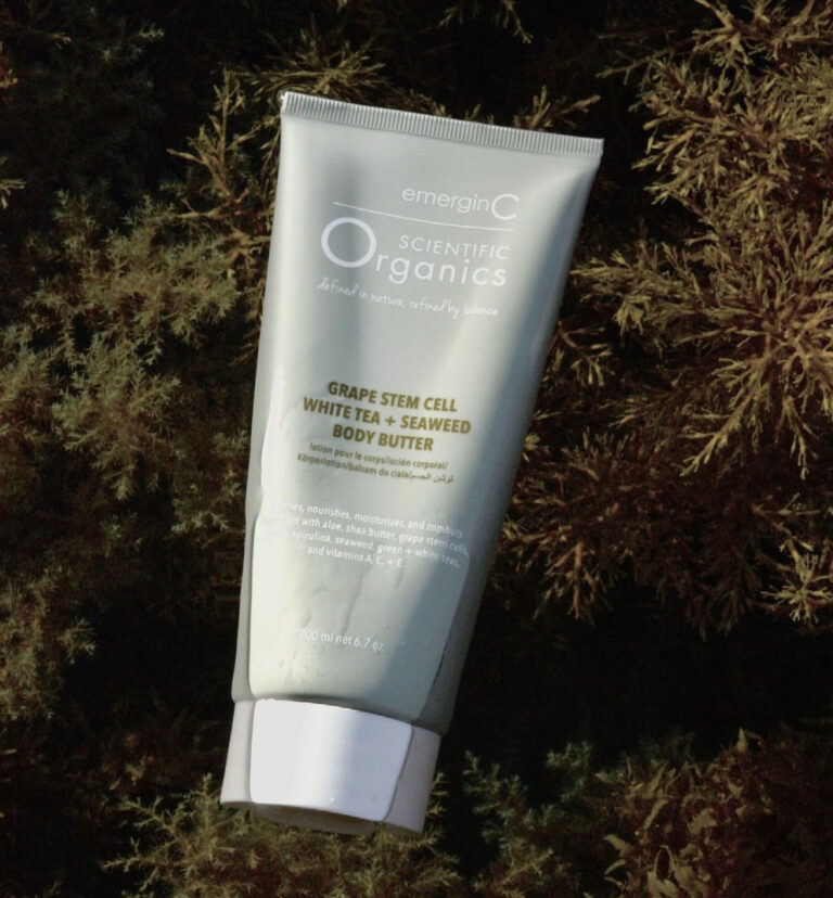 A tube of professional luxury skin care, emerginC Scientific Organics Grape Stem Cell White Tea + Seaweed Body Butter, is placed on a bed of brown seaweed. The grayish-green tube with white text and cap stands out against the natural background.