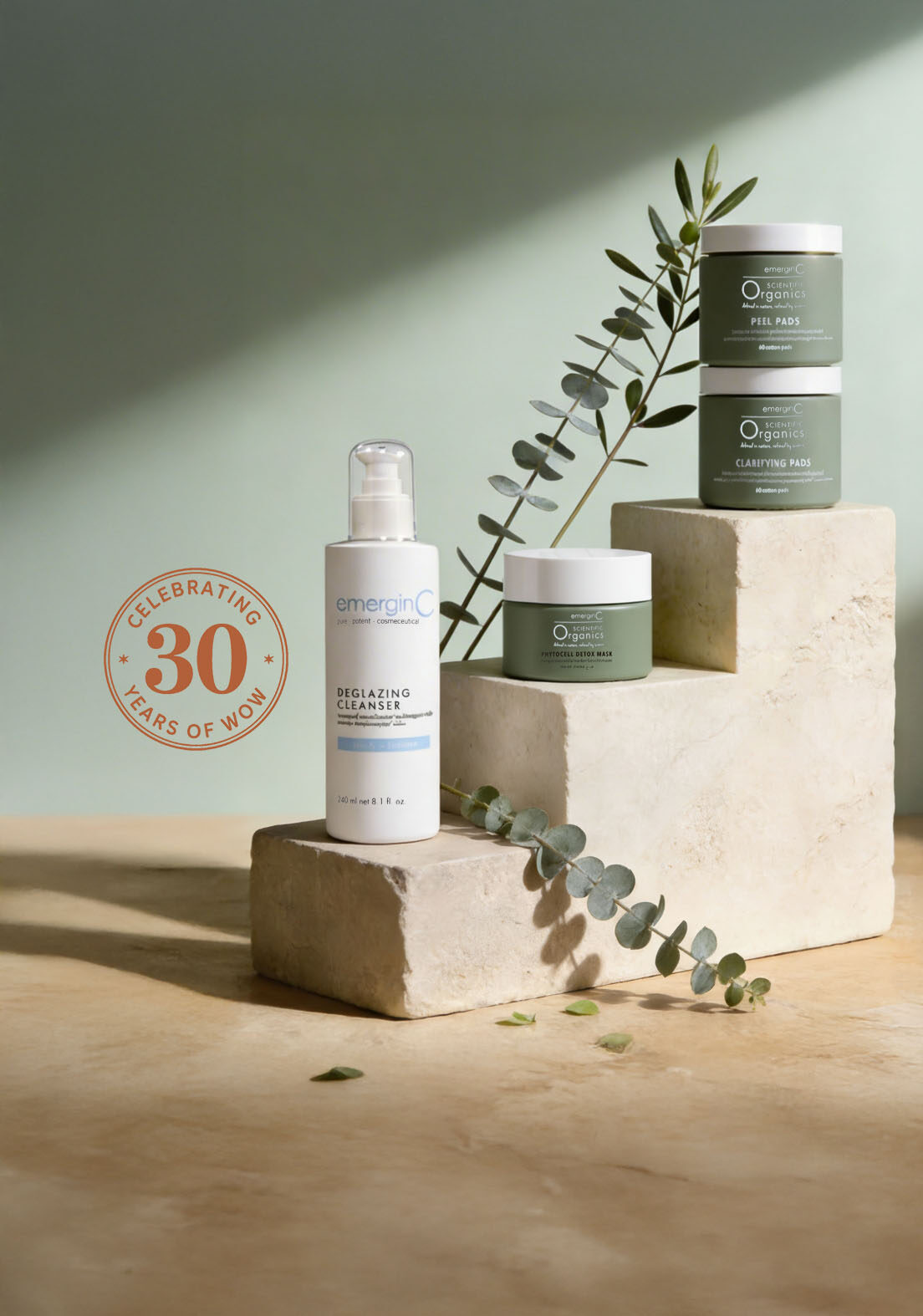 A collection of professional luxury skin care products displayed on stone blocks with eucalyptus leaves. A circular badge reads “Celebrating 30 Years of WOW!” Cleanser and jars with green labels are arranged in soft natural light.