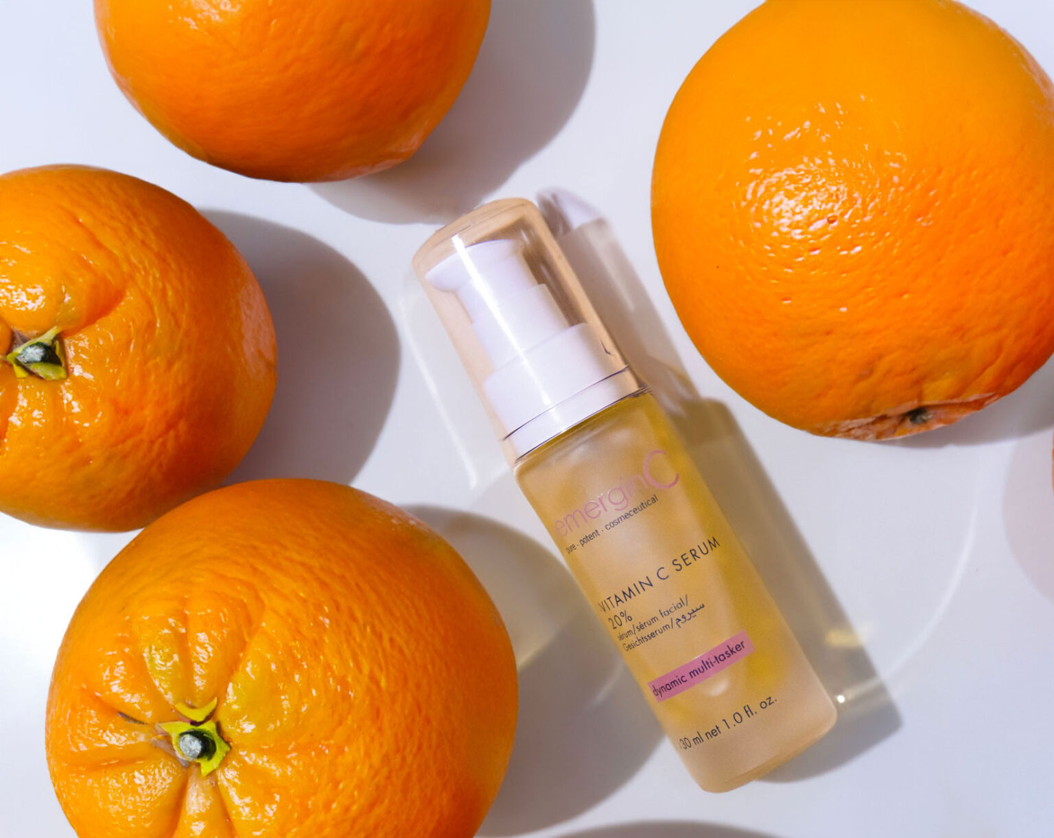 A bottle of vitamin C serum with a pump sits on a white surface surrounded by four fresh oranges. Perfect for spa skin care routines, the labeled serum bottle is positioned diagonally among the oranges.