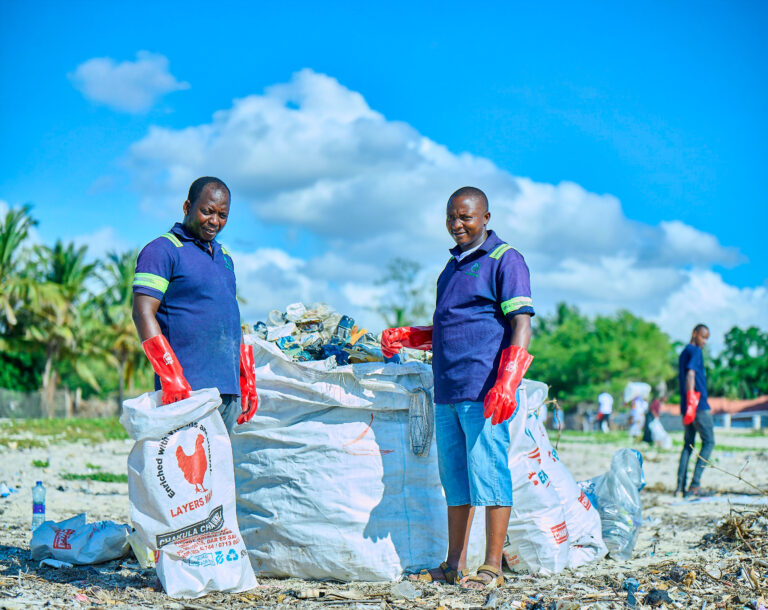 Two people wearing blue shirts, shorts, and red gloves stand on a beach next to large white bags filled with collected trash, helping clean up litter under a bright blue sky—perfect for those who appreciate nature as much as top skin care lines for spas.
